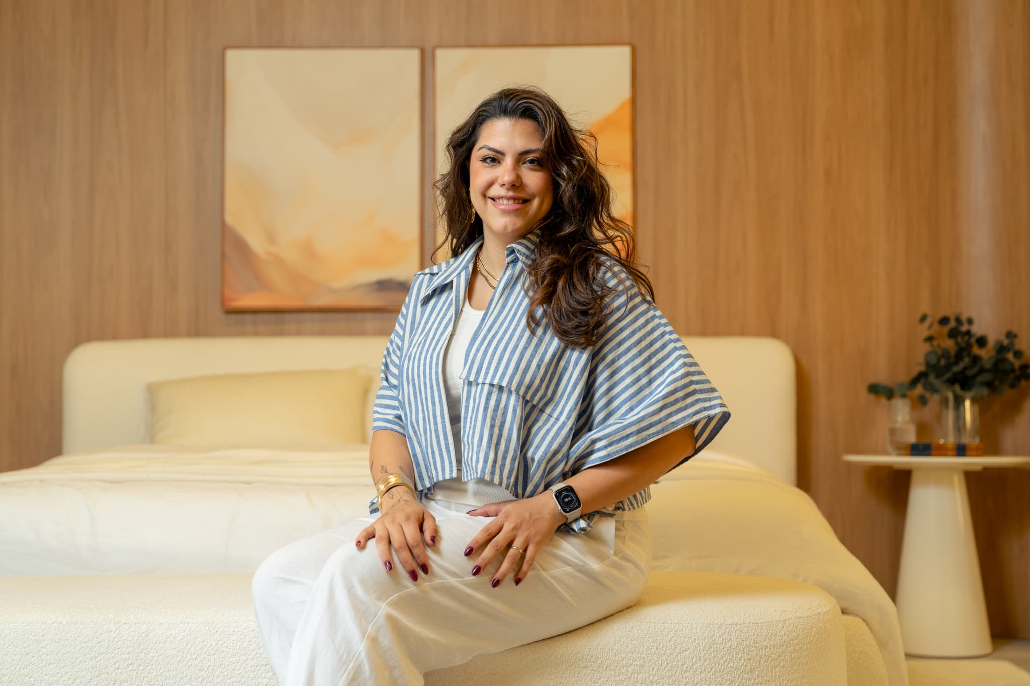 Daniely Périco, sócia da Studio Side, sorrindo sentada na beira de uma cama clara, diante de um painel de madeira e de um quadro em tons de terra