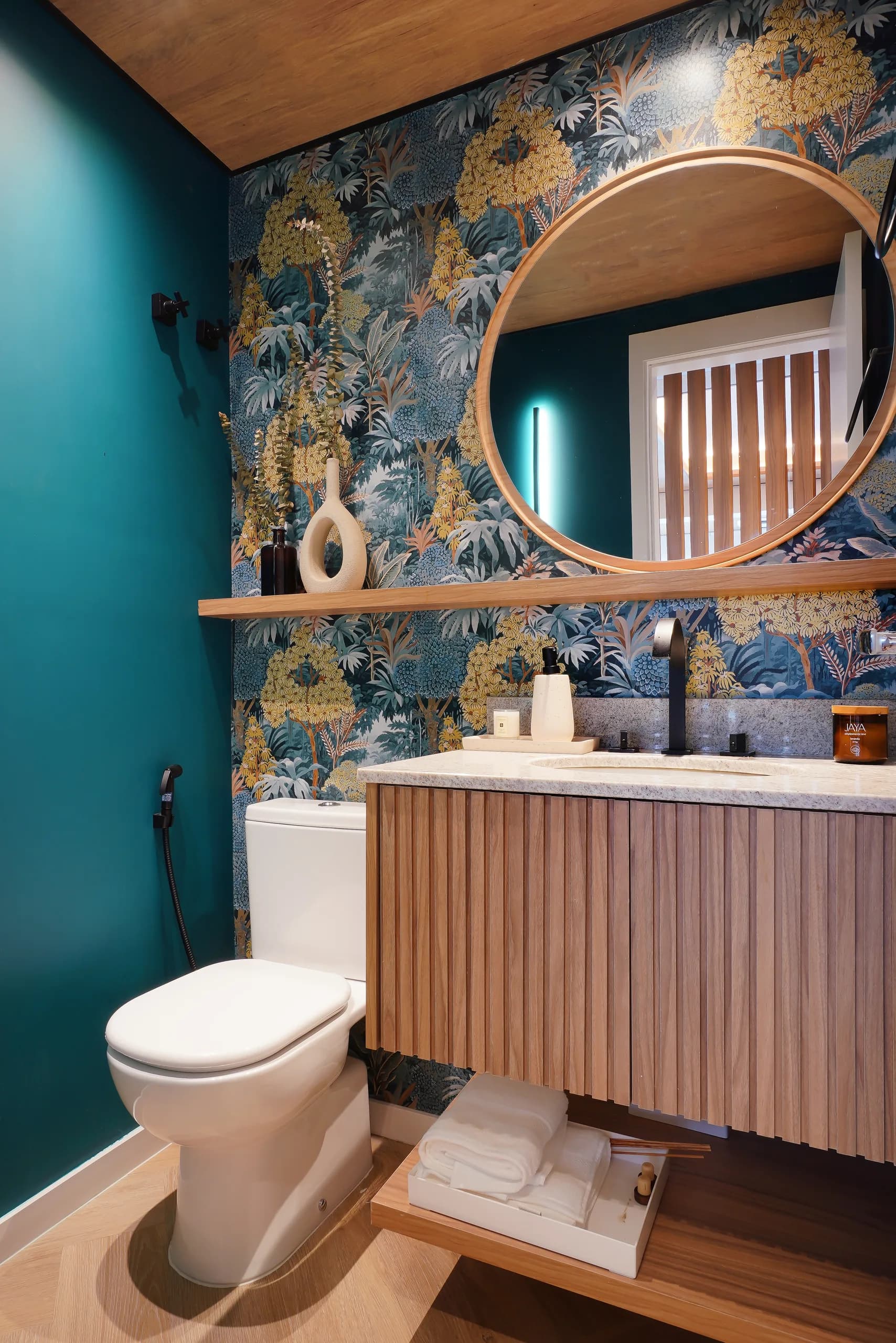 Lavabo com papel de parede floral, espelho redondo, marcenaria ripada e parede verde