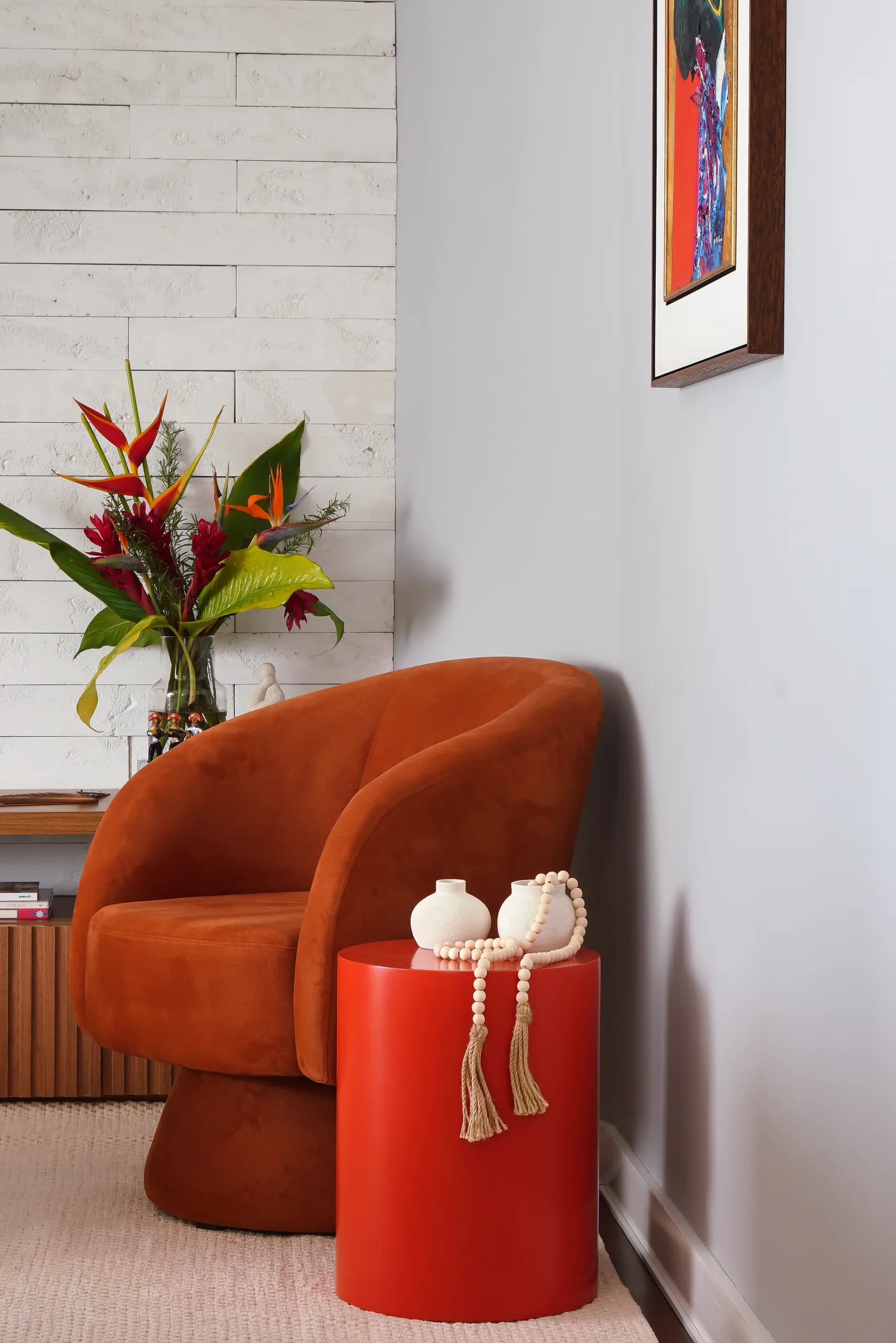 Poltrona estofada em terracota ao lado de mesa lateral vermelha, com flores e parede de tijolo branco