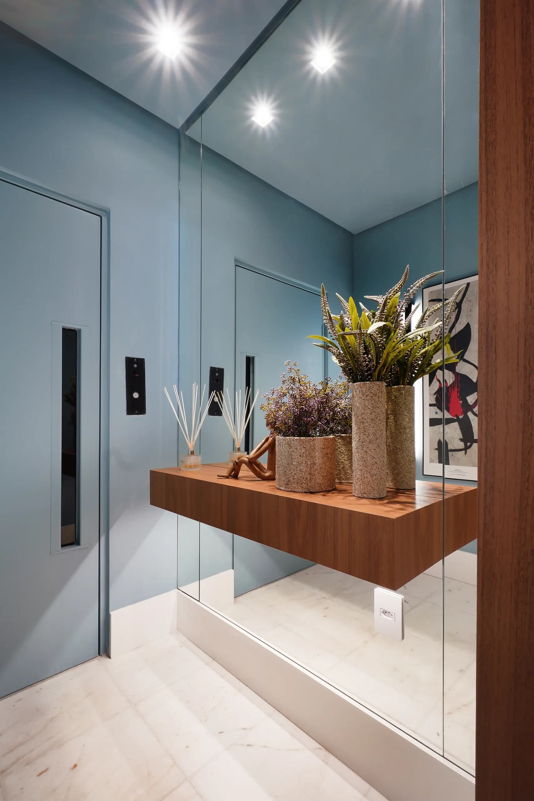 Hall de entrada azul com espelho, aparador suspenso de madeira com vasos e plantas e piso de mármore claro