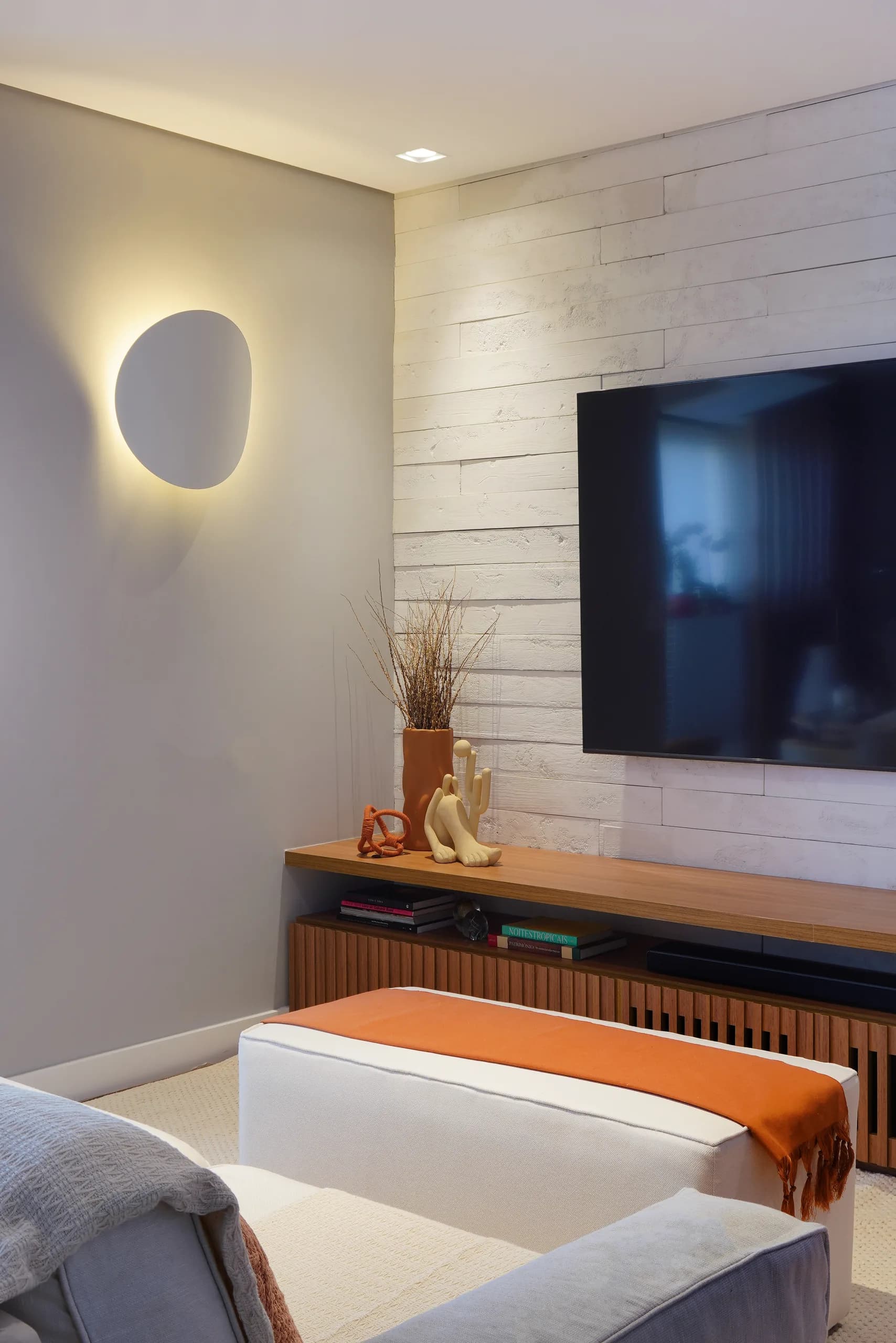 Arandela de formato orgânico iluminando a parede ao lado da TV, com rack de madeira ripada e pufe com manta laranja